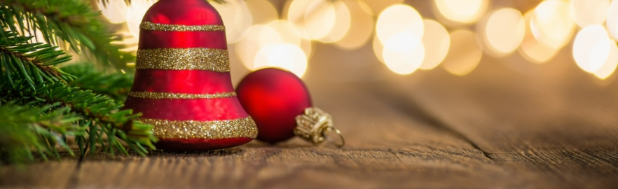 Świąteczna kompozycja z czerwoną bombką w kształcie dzwonka ze złotym brokatem i gałązkami jodły, leżąca na rustykalnym drewnianym stole. W tle rozmyte, ciepłe światła.