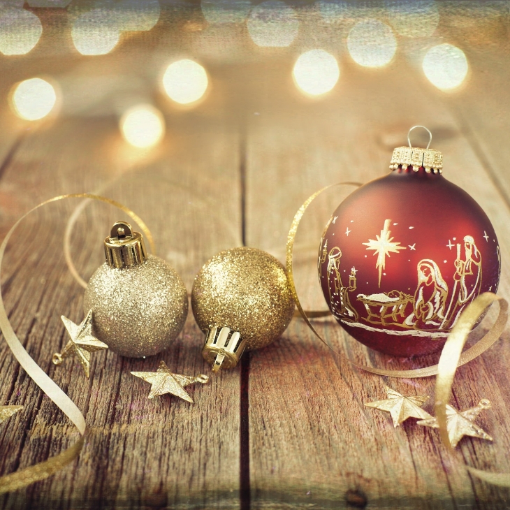 Czerwona bombka z motywem szopki bożonarodzeniowej i dwie mniejsze, złote, brokatowe bombki, ułożone na rustykalnym drewnianym stole w otoczeniu złotych wstążek i gwiazdek.