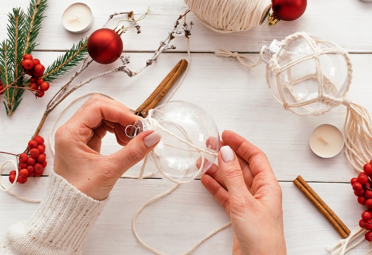 Ręce oplecione sznurkiem na przezroczystej bombce, w tle świąteczne dekoracje (cynamon, czerwone koraliki).
