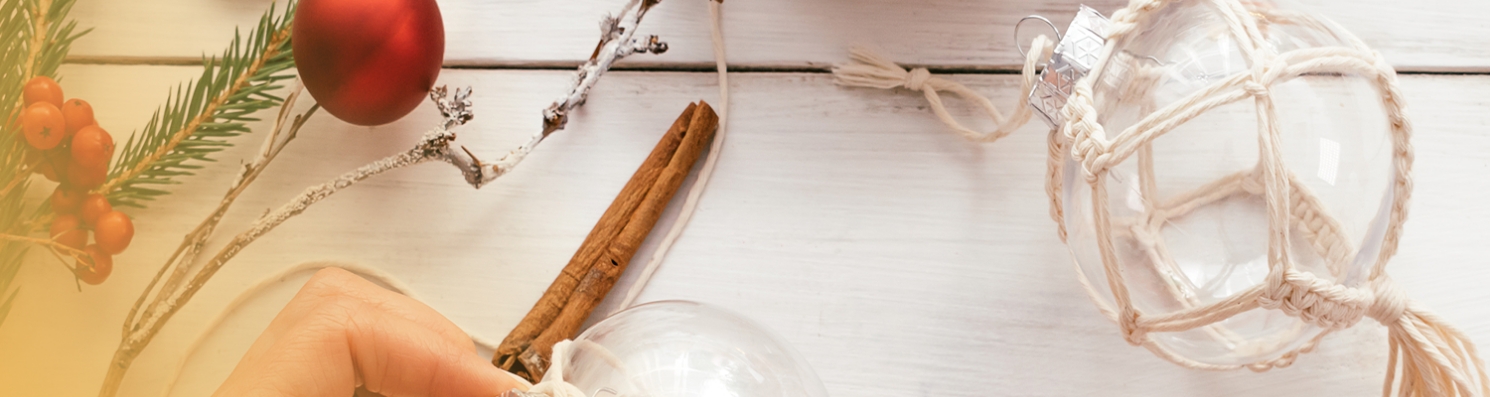 Tworzenie naturalnych ozdób świątecznych – Ręka dekorująca szklaną bombkę sznurkiem. Na białym stole gałązki jodły, cynamon i czerwone jagody.