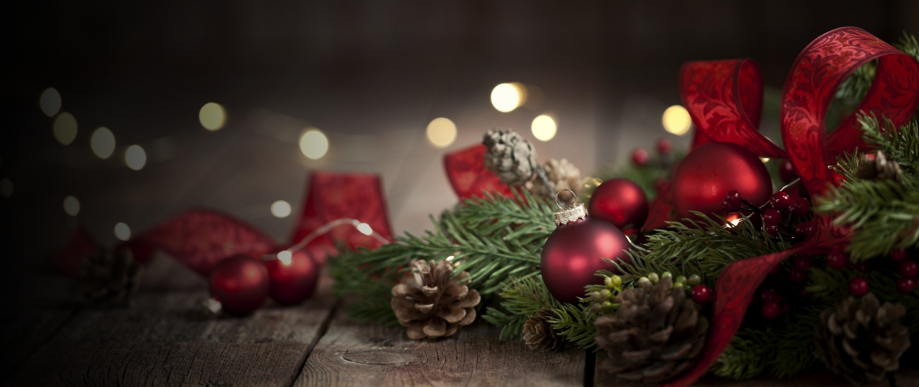 Świąteczna dekoracja z czerwonymi bombkami, szyszkami, zielonymi gałązkami i czerwoną wstążką na drewnianym stole.